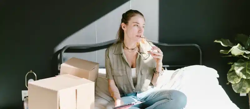 A woman sits with tablet and coffee, organizing her home-based e-commerce setup with boxes.