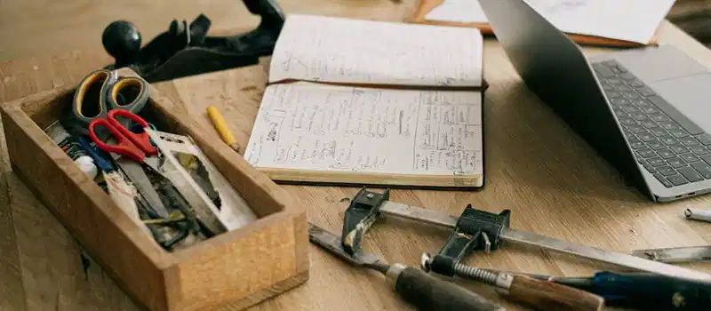 A creative woodworking workspace with tools, notebook, and laptop on a wooden table.