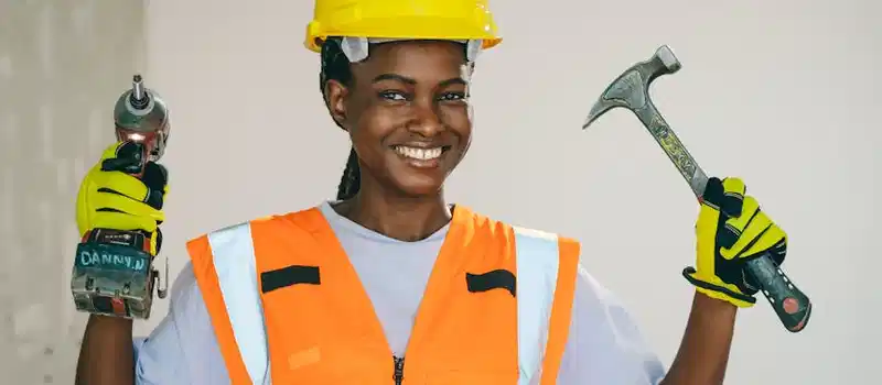 Smiling female engineer wearing safety gear, holding a drill and hammer confidently.