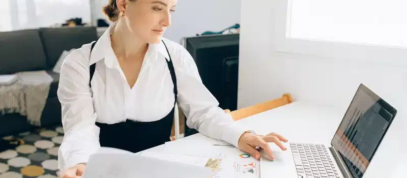 Woman at desk reviewing business reports on laptop, holding papers in a modern office setting.