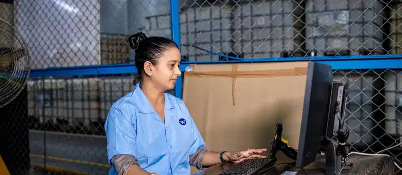 A woman in blue uniform operates a computer inside a factory warehouse, focusing on her task.