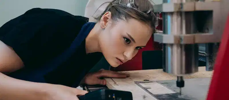 Woman closely inspecting machinery in a workshop wearing protective goggles.