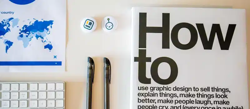 Flat lay of a creative workspace with 'How To' book, chart, pens, and keyboard in minimalist style.