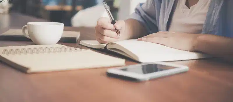 A woman writes in a notebook at a café table with a coffee and smartphone nearby.