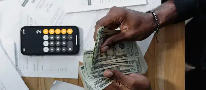 Person counting dollar bills over documents with a smartphone calculator on the desk.