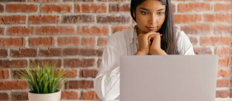 Young woman deeply focused on her laptop studying indoors against a brick wall backdrop.