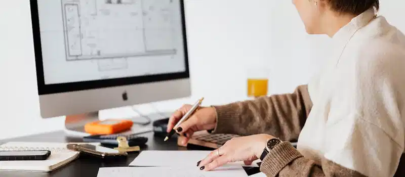 Woman designing architectural blueprints using desktop computer in a modern office setting.