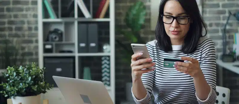 Professional woman using phone and credit card for online transaction at desk in office.