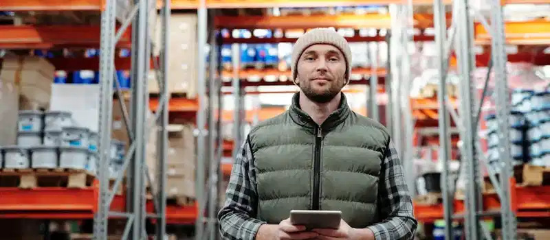 A warehouse worker wearing a beanie uses a tablet to manage inventory in a storeroom with shelves.