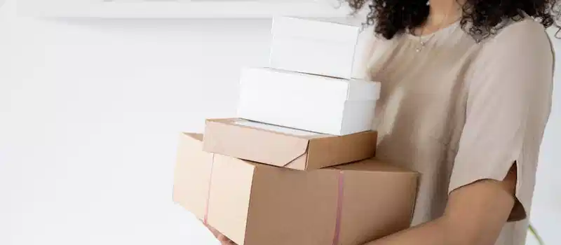 Woman with curly hair carrying stack of packaging boxes in a bright room.