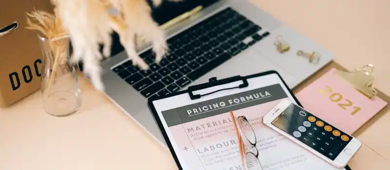 Organized office desk with pricing formula document, laptop, and smartphone calculator.