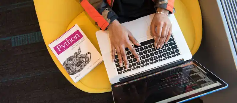 A developer typing code on a laptop with a Python book beside in an office.