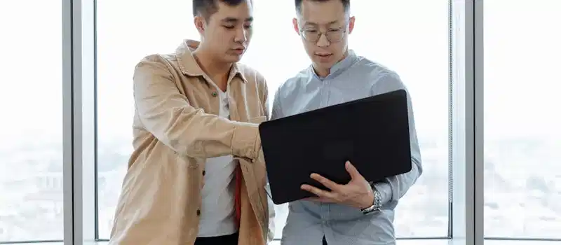 Two young professionals in an office setting, collaborating on a project using a laptop.