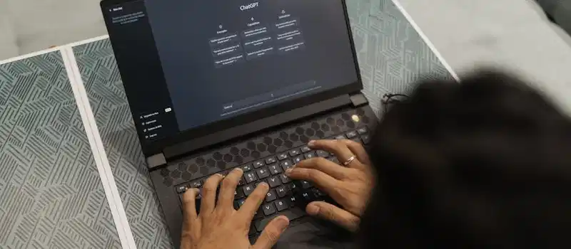 Close-up of hands typing on a laptop displaying ChatGPT interface indoors.