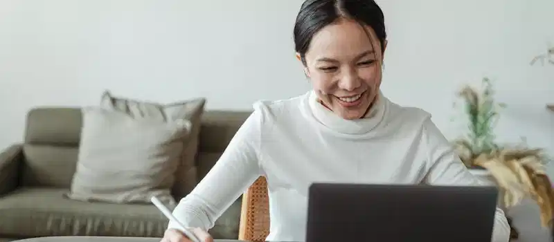 A cheerful woman uses a laptop and tablet for a video call, working remotely in a cozy living room.