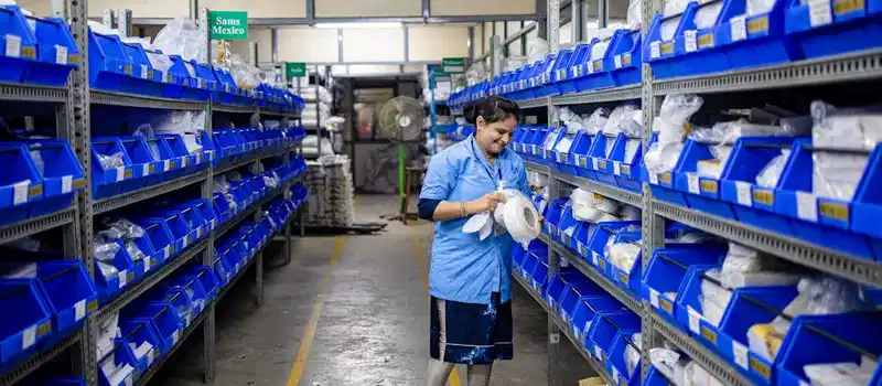 A woman organizing inventory in a warehouse aisle, surrounded by shelves and blue bins.