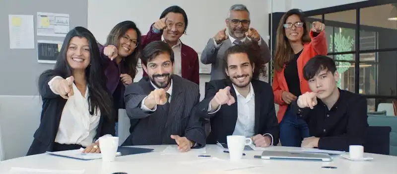 A diverse group of colleagues smiling and pointing in a modern office setting, promoting teamwork.