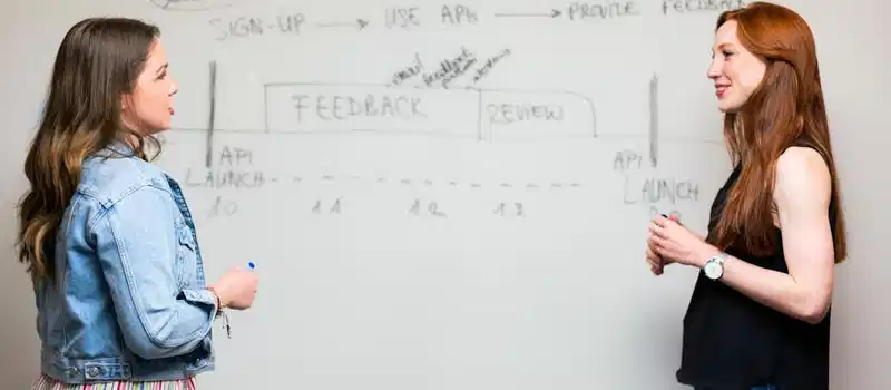 Two women engaging in a discussion about API development processes at a whiteboard.