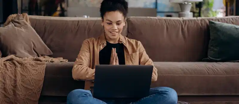 A smiling young woman sits cross-legged on a pouf, using her laptop in a cozy home setting.