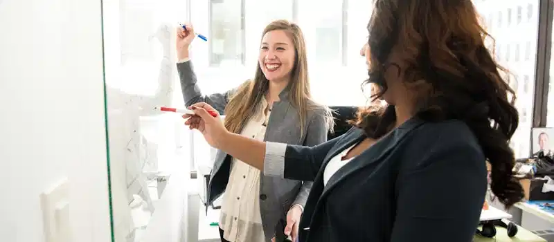 Two professional women discussing ideas on a whiteboard in a modern office setting.