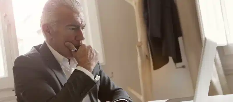 Elderly businessman in a suit thoughtfully working on a laptop at his desk.