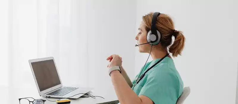 A female doctor using a laptop for an online consultation, wearing a headset in a bright office.