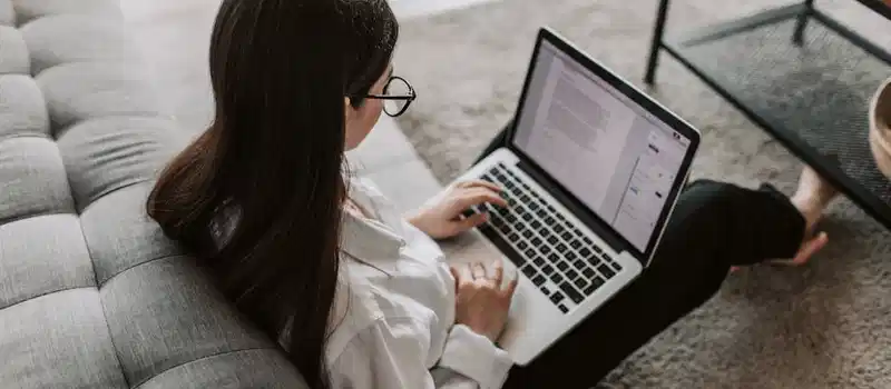 Woman working remotely with a laptop on the floor next to a sofa, enjoying comfortable home office setup.