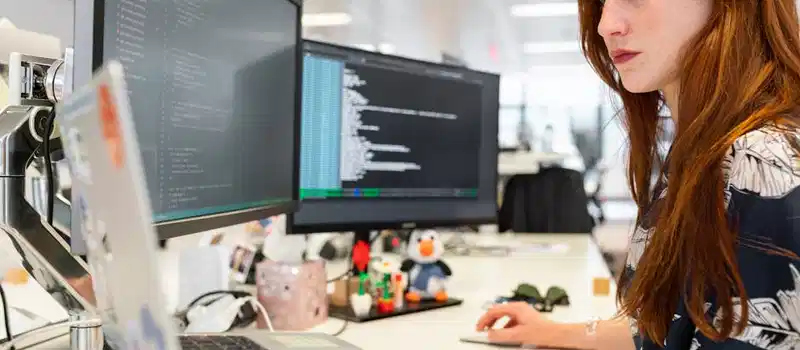 A focused female software engineer coding on dual monitors in a modern office.