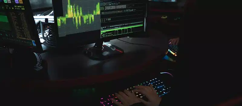 A cybersecurity expert in a dimly lit room is typing on a colorful keyboard with multiple screens displaying data.