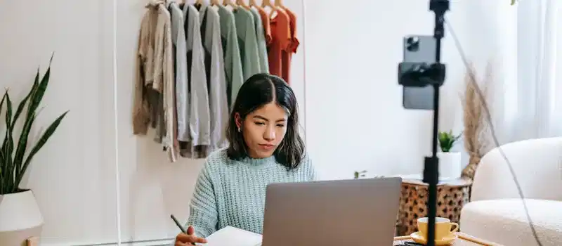 Focused young woman working on laptop in home office, surrounded by clothing rack and camera setup.