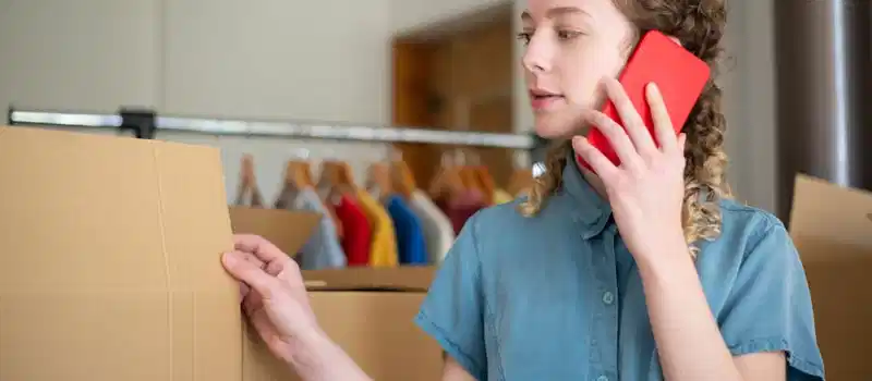 Young woman in blue shirt using phone while packing cardboard boxes indoors.