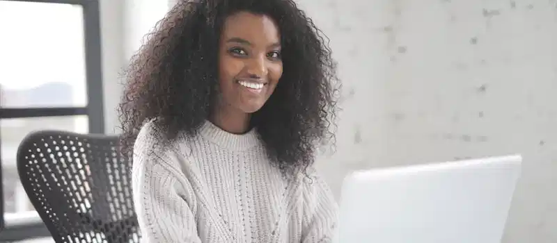 Smiling woman with curly hair using a laptop in a modern workspace, showcasing remote work confidence.