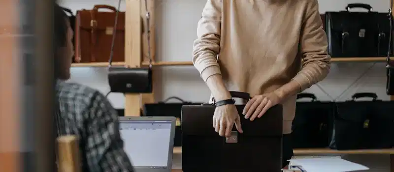 Young artisan selecting leather briefcase in workshop with laptop and numerous bags displayed in the background.
