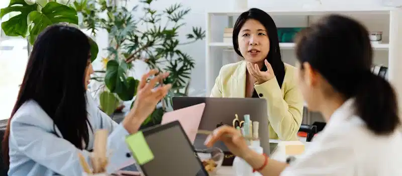 Three businesswomen engaged in discussion over laptops in a modern office setting.