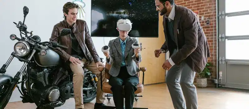 Two men assist a seated colleague with a virtual reality headset in a stylish, brick-walled office.
