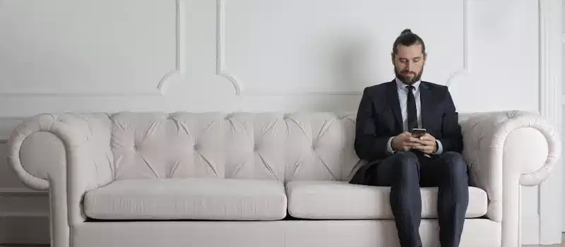 A businessman in a suit sits on a stylish white sofa using a smartphone indoors.