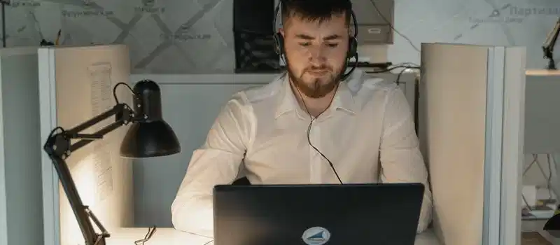 A bearded call center agent wearing headphones, focused on his laptop at work in a modern office.