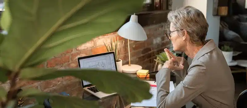 Senior woman in a modern home office focusing on work at a laptop next to a brick wall.