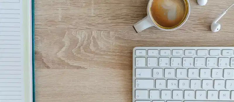 Flat lay of a workspace featuring coffee, a keyboard, and a notebook on a wooden desk.