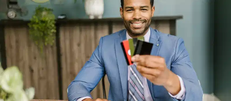 Smiling African American man in a suit holding credit cards at a business desk.