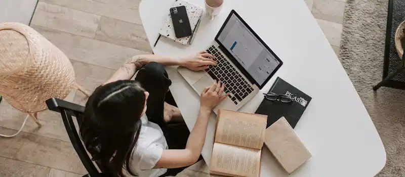 Overhead view of a woman using a laptop at a home desk, surrounded by books, a phone, and a cup.
