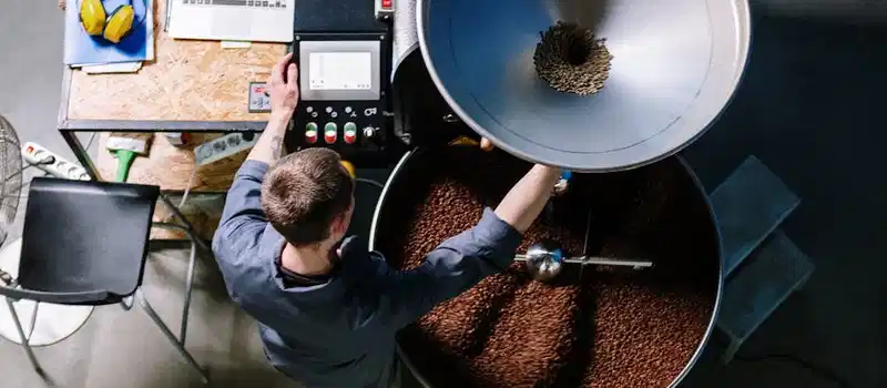 A worker in a coffee roastery operating equipment, processing fresh beans in an industrial setting.