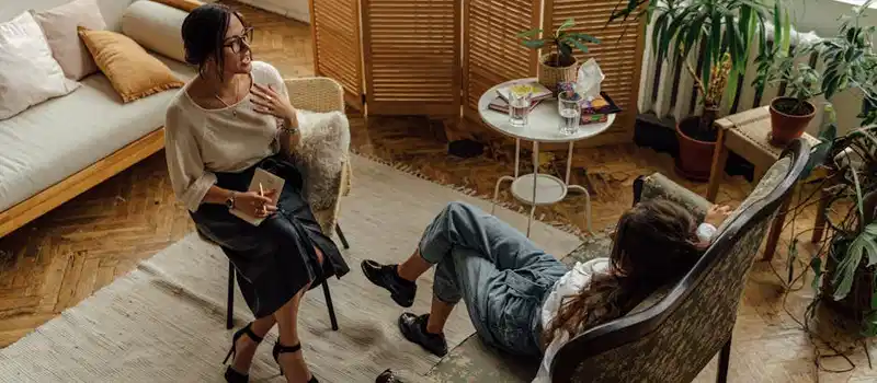 Two women engaged in a psychotherapy session in a warm, inviting interior with plants and natural lighting.