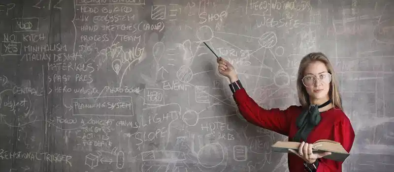 Serious female teacher wearing old fashioned dress and eyeglasses standing with book while pointing at chalkboard with schemes and looking at camera
