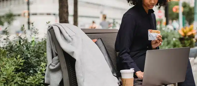 Young woman sitting on a bench using a laptop and enjoying a coffee and sandwich outdoors.