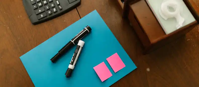 Top view of office supplies including a calculator, markers, and sticky notes on a wooden desk.