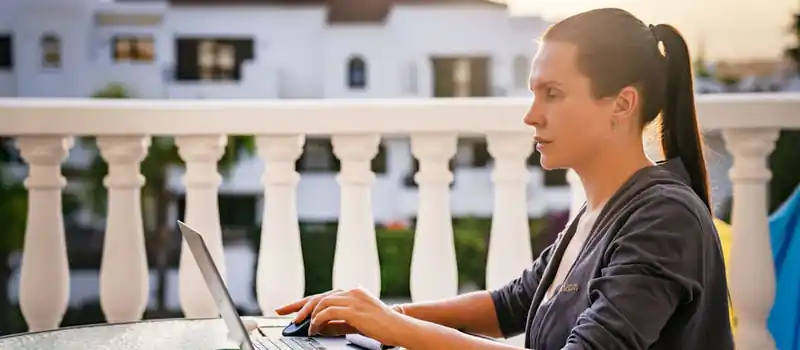 A focused woman works on her laptop outdoors on a sunny balcony, capturing a serene morning work vibe.
