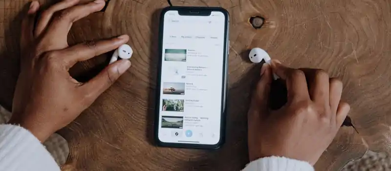 Hands interacting with smartphone and earbuds on a rustic wooden table, highlighting modern technology.