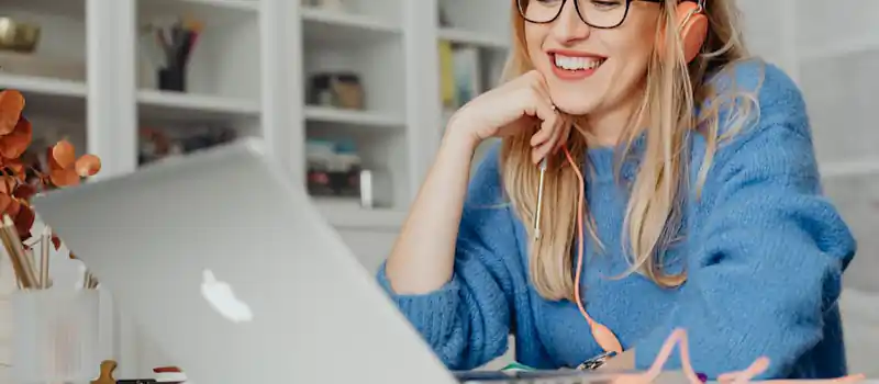 Smiling woman wearing headphones and glasses, working on a laptop in a bright indoor setting.