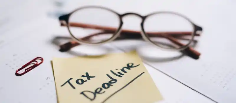 A close-up shot of a sticky note with 'Tax Deadline' written on it, alongside eyeglasses and a calendar.